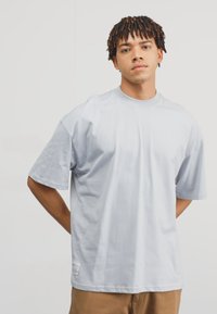 Lys grå oversized t-shirt i blødt stof, med rund halsudskæring og korte ærmer samt et lille mærke nederst til venstre.