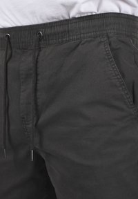 Close-up van zwarte joggingsbroek met zichtbare tailleband, trekkoorden en zijzak op een persoon die een wit shirt draagt.