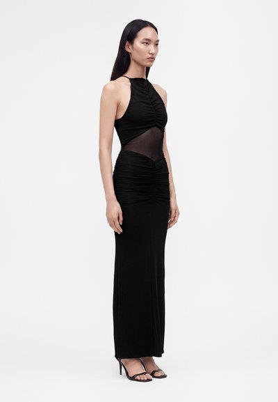 Femme aux longs cheveux foncés portant une robe de soirée noire sans manches avec des détails froncés et une partie médiane transparente, debout dans des sandales noires à talons hauts.