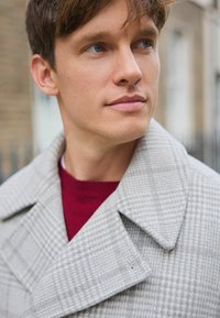 Harmaa houndstooth-villakangas takki suurilla läpipainolevyillä, päällä burgundinvärinen villapaita. Lähikuva kaulan alueesta ja ylärinnasta. Huomaamaton tekstuurin yksityiskohta näkyvissä.