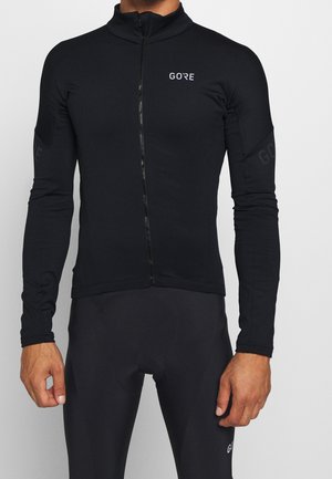 Uomo che indossa una giacca sportiva nera con zip a maniche lunghe e pantaloni sportivi neri abbinati con il logo "GORE" sul petto e sulla gamba.