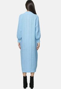 Kvinde stående med ryggen mod, iført en lang, lyseblå strikket kjole med ballonærmer og sorte støvler med høje hæle.