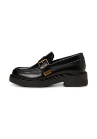 Mocassin en cuir noir avec une semelle en wedge épaisse, présentant une boucle en laiton et des coutures décoratives le long du bord supérieur.