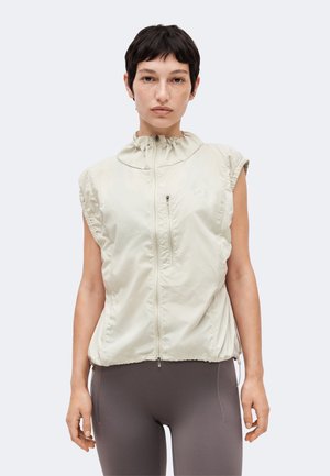 Kobieta z krótkimi ciemnymi włosami, ubrana w bezrękawnik jasnobeżowy zapinany na zamek, na ciemnoszarych legginsach, stojąca na jednolitym tle.