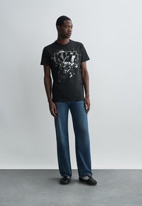 Čierne bavlnené tričko s potlačou KISS, spárované s voľnými modrými džínsami. Model stojí na šedom podlahe pred svetlou stenou.