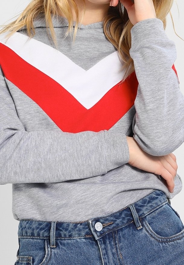 Szara bluza z kapturem z czerwono-białym wzorem w kształcie litery V na piersi, z prążkowanymi mankietami i fakturowanym materiałem. Noszona z dżinsami.