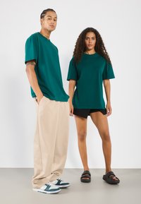 Dva modely v ateliéru nosí tyrkysové oversize trička, béžové široké kalhoty, černé šortky a pohodlné tenisky.