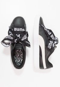 Černé kožené tenisky s širokými černobílými značkovými tkaničkami, bílými gumovými podrážkami a texturovanou vnitřní stélkou s černobílým vzorem.