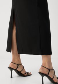 Black long dress with a side slit, paired with black strappy high-heel sandals. Smooth fabric with a tailored fit and clean hem.