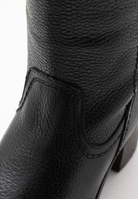 Primer plano de una bota de cuero negro texturizado con costuras detalladas y un tacón cuadrado sobre un fondo blanco.