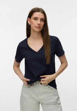 Young woman with long brown hair wearing a navy V-neck T-shirt and light gray high-waisted pants, adjusting her shirt at waist level.