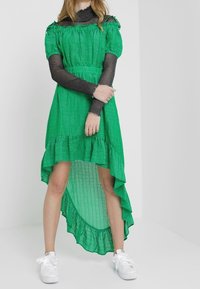 Vaatteet vihreällä kuviolla, jossa on korkea matala helma, röyhelökoristeet ja lyhyet puffihihat, kerroksittain mustan pitkähihasen paidan päällä ja yhdistettynä valkoisiin tennareihin.