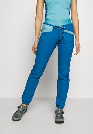 Vrouw die blauwe sportbroek met trekkoord, lichtblauw shirt en grijze wandelschoenen draagt, staand tegen een witte achtergrond.