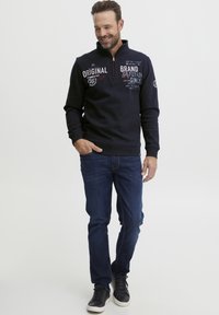 Felpa mezza zip di un blu navy scuro con stampe di testo bianche e rosse, abbinata a jeans blu scuro e sneakers nere con suola bianca.