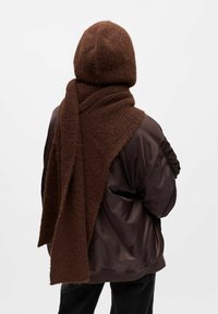 Bruine gebreide muts en sjaal gecombineerd met een bruine, glanzende pufferjack. De sjaal heeft een gestructureerd patroon en hangt losjes.