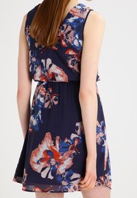 Robe florale marine avec des fleurs rouges, bleues et blanches vibrantes ; design sans manches et taille froncée, fabriquée en tissu léger.