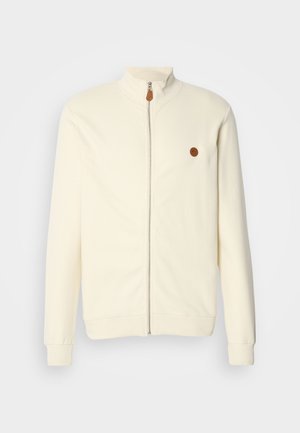 Jachetă crem cu fermoar, cu guler înalt, textură ribbed și un mic patch cu logo maro pe pieptul stâng. Are mâneci lungi și manșete elastice.