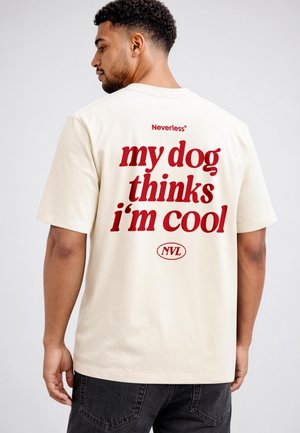 Mann mit kurzen lockigen Haaren steht seitlich, trägt ein cremefarbenes T-Shirt mit rotem Text auf dem Rücken: „Mein Hund findet mich cool“.