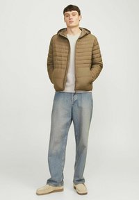 Gepolsterte olivgrüne Jacke mit Kapuze, ausgestattet mit einem Reißverschluss und horizontaler Steppung; kombiniert mit hellblauen, lässig geschnittenen Jeans und beigen Schuhen.