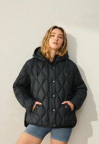 Giacca nera imbottita con cappuccio, caratterizzata da un motivo a rombi, bottoni a pressione e tasche laterali. Indossata sopra pantaloni shorts blu.