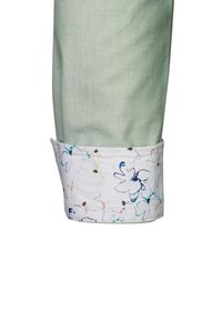 Tessuto verde chiaro con una superficie liscia, caratterizzato da un polsino bianco a motivo floreale con accenti blu e gialli, che mostra fiori delicati.
