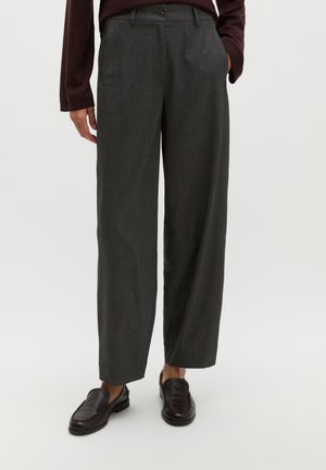 Person trägt dunkelgraue, weit geschnittene Hose, dunkelbraune Loafers und ein dunkles Langarmshirt, eine Hand in der Tasche.