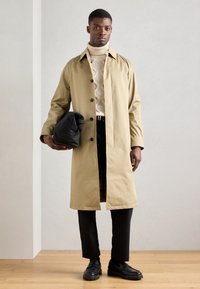 Beige trenchcoat met zwarte knopen, gedragen over een crèmekleurige kabelbreiselsweater, gecombineerd met zwarte broek en schoenen, en houdt een zwarte tas vast.