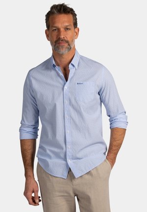 Man met grijze baard, gekleed in een lichtblauw overhemd met opgerolde mouwen en beige broek, staand tegen een effen achtergrond.