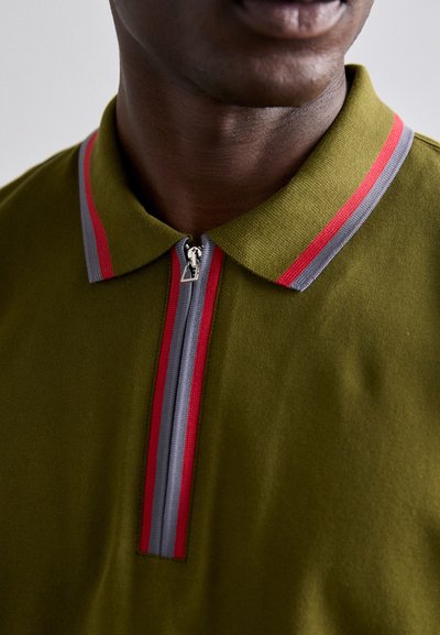 Polo de color verde oliva con cuello con cremallera, con acentos a rayas rojas y grises en el cuello y la tapeta de la cremallera. Material de mezcla de algodón.