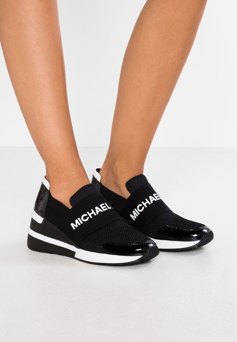 Schwarze Slip-On-Sneaker mit Mesh-Stoff und Lacklederakzenten. Sie verfügen über einen weißen Streifen mit dem Markennamen auf der Vorderseite und eine Keilsohle.