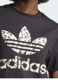 Svart t-shirt med stort Adidas-logotyp som har ett ljust leopardmönster, bärs av en person med mörk hud och lockigt hår.