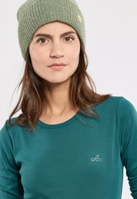 Groene geribbelde beanie met een klein logo; teal kleurige longsleeve top die een aansluitend ontwerp heeft en glimmende logo-details op de borst.