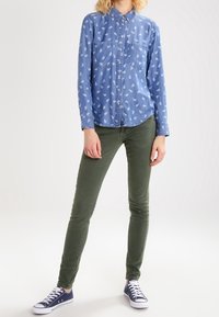 Camisa de botones azul claro con patrón de hojas blancas, dos bolsillos en el pecho; combinada con jeans skinny verdes y zapatillas azul marino.