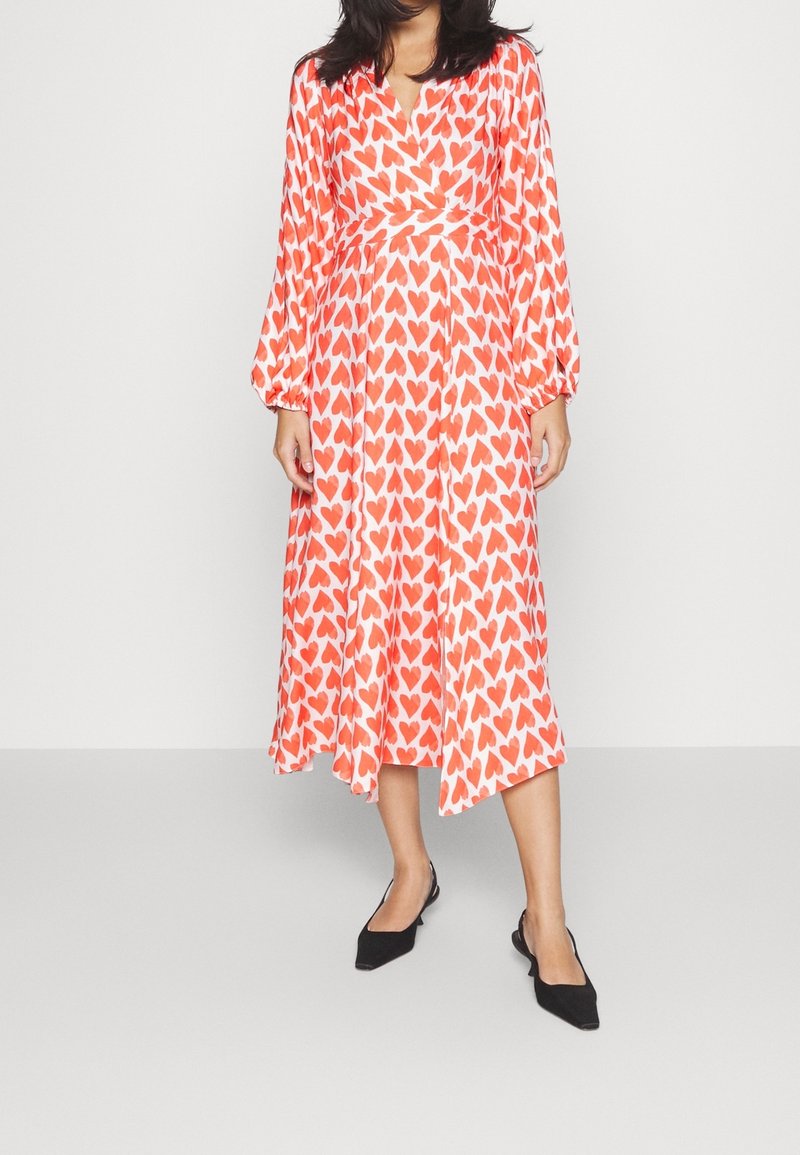 Robe enroulée rouge et blanche avec de longues manches bouffantes, présentant un motif en forme de cœur. Tissu lisse, longueur genoux avec un ourlet arrondi. Ballerines noires à bouts pointus.