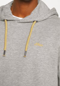 Sudadera con capucha gris hecha de tela suave con acabado texturizado, con cordones amarillos y terminales plateados. Pequeño logo bordado de "s.Oliver".
