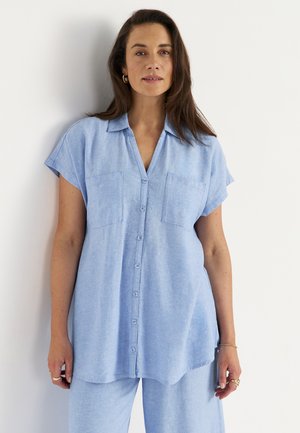 Kobieta o długich ciemnych włosach, ubrana w jasnoniebieską koszulę na guziki z krótkim rękawem i dopasowane spodnie, stojąca przy białej ścianie.