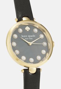 Guldfärgad klocka med svart läderarmband. Grå urtavla med en spade-logotyp, pärlursmarkeringar och guldfärgade visare. Klassisk rund form.