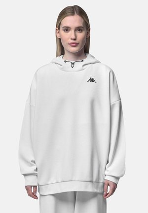 Jeune femme portant un sweat à capuche blanc oversized avec un logo noir sur la poitrine, debout devant un fond clair uni.