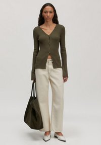 Olivgrüner gerippter Cardigan mit Knöpfen, kombiniert mit cremefarbenen weit geschnittenen Hosen. Die Accessoires umfassen eine dunkle Tragetasche und weiße spitze Ballerinas.
