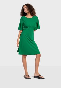 Femme aux cheveux bouclés portant une robe verte mi-longue avec des manches volantées et des tongs à plateforme noires, debout devant un fond uni.