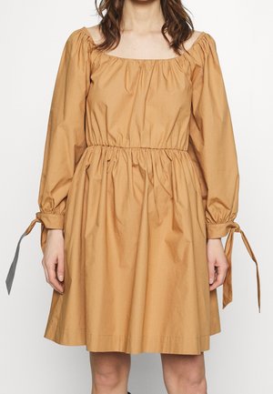 Kvinde iført en knælang tan-farvet kjole med lange pufærmer og bundne manchetter, med en elastisk indsamlet talje og firkantet halsudskæring.