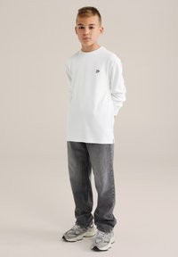 Vit långärmad t-shirt med en liten svart logga, paired med mörkgrå jeans och gråa sportskor med svarta och ljusgröna accenter.
