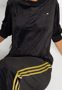 Schwarzes Velour-Oberteil mit lockerer Passform und einem dezenten geprägten Muster. Mit goldenem Logo und schwarzen gerippten Bündchen. Kombiniert mit schwarzen Hosen und goldenen Streifen.