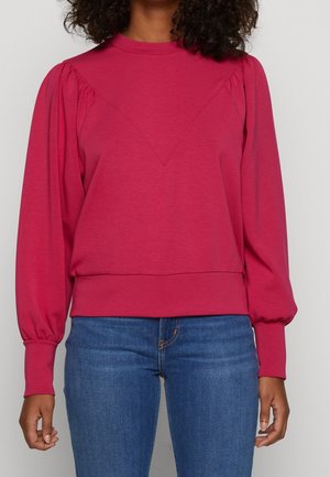 Kobieta ubrana w długą bluzkę w kolorze malinowym z bufkami na ramionach i dopasowanymi mankietami, w połączeniu z niebieskimi dżinsami.