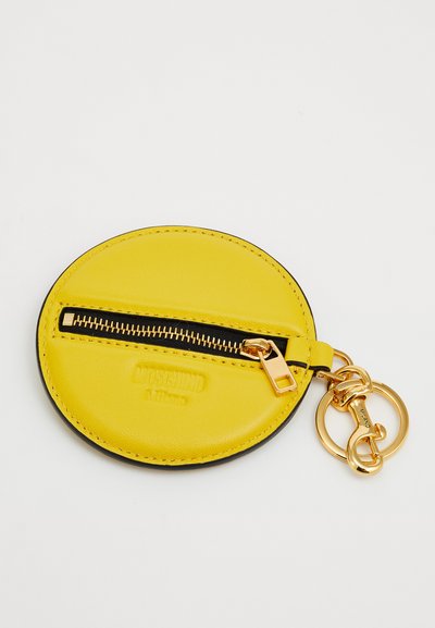 Στρογγυλό κίτρινο δερμάτινο πορτοφολάκι για κέρματα με μαύρο φερμουάρ και χρυσούς κρίκους μπρελόκ, με ανάγλυφο το εμπορικό σήμα "Moschino Milano" στην πρόσοψη.