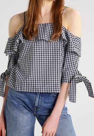 Kobieta w czarno-białej bluzce w kratę gingham z odkrytymi ramionami, falbankami i wiązanymi rękawami 3/4, połączona z niebieskimi jeansami.