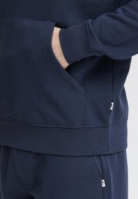Sudadera azul marino con un bolsillo tipo canguro en la parte frontal, puños acanalados y dobladillo. Textura de tela suave con acentos de marca mínimos.