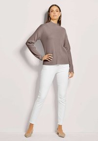 Brauner Strickpullover mit Mock-Neck, seitlichen Schlitzen und gerippten Ärmeln, kombiniert mit weißen, eng anliegenden Jeans und hellbeigen Absätzen.