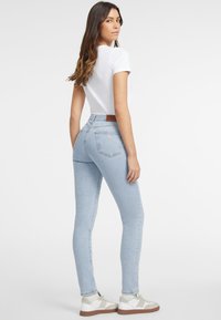 Jeans skinny blu chiaro con vita alta, caratterizzati da una patch in pelle marrone sul retro, abbinati a una maglietta bianca e sneakers.