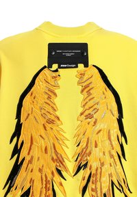 Sudadera amarilla con intrincadas alas bordadas en oro y negro en la parte posterior, con una etiqueta negra y detalle de cierre a presión.